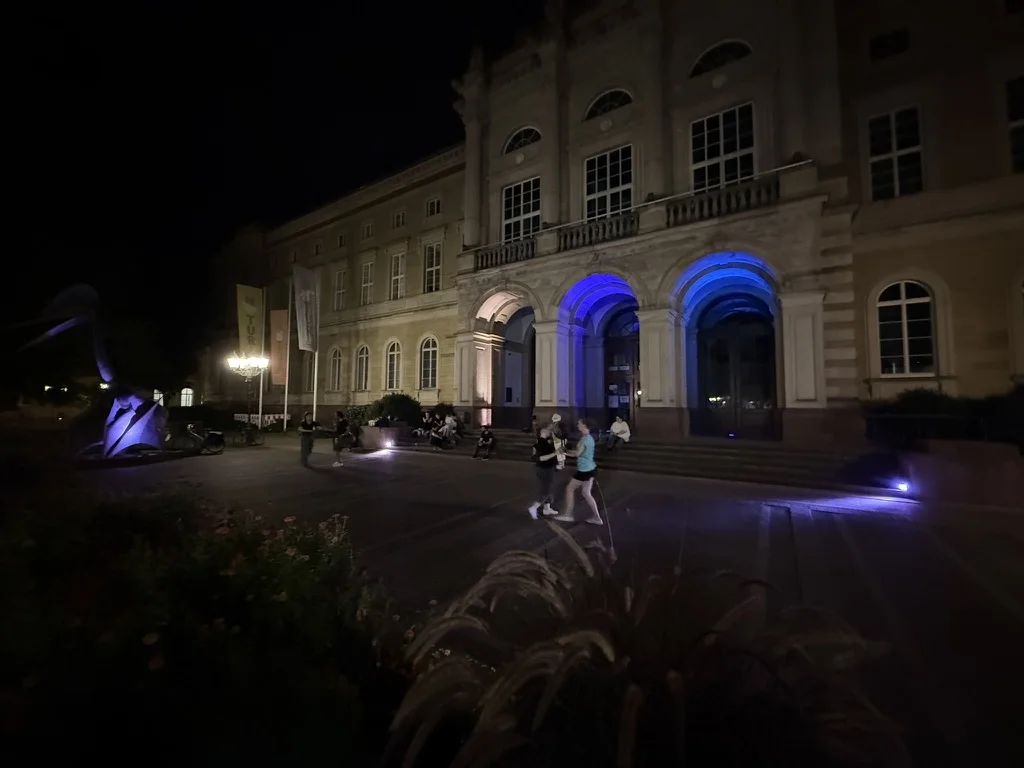 Tanzende vor dem Naturkundemuseum bei Nacht, dessen Eingangsbögen blau und violett beleuchtet sind