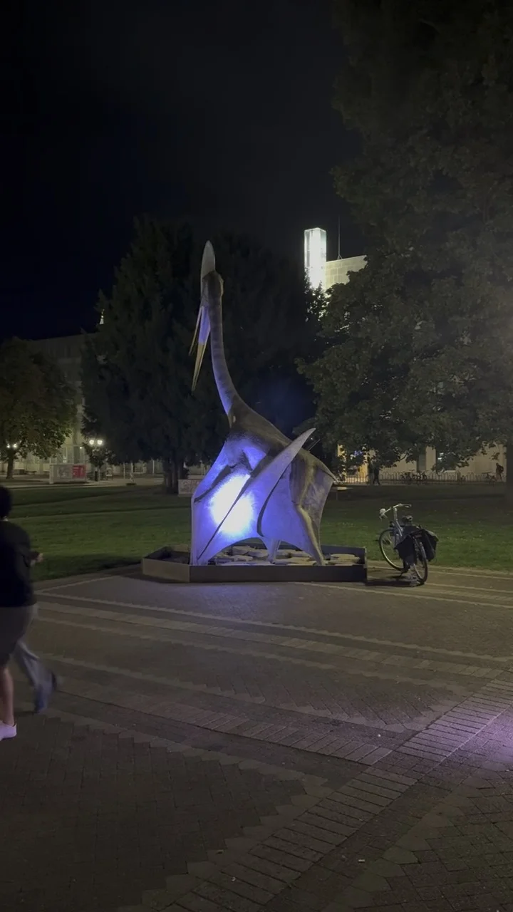 Blau angestrahlte Flugsaurier-Skulptur vor dem Museum, daneben steht ein Fahrrad